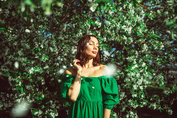 A young beautiful girl stands in the park under a blossoming apple tree, wearing a beautiful bright green dress.