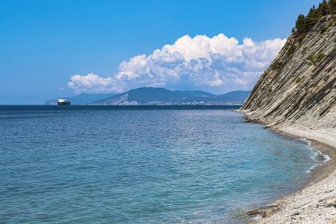 Güzel yaz manzarası, bulutlu parlak mavi gökyüzü, ağaçlı dik uçurumlar, taştan vahşi plaj ve ufuktaki Novorossiysk şehri manzarası. Rusya, Gelendzhik, Karadeniz kıyısı