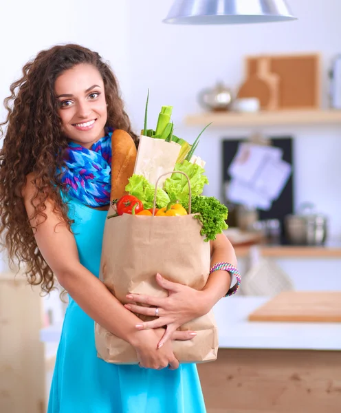 Ung kvinde med indkøbspose med grøntsager stående i køkkenet. - Stock-foto