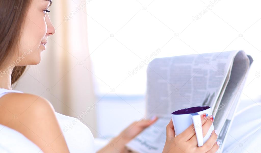 A pretty young woman reading the newspaper in bed and enjoying a cup of tea