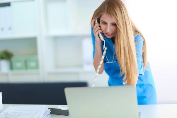 stock image Young woman doctor in white coat at computer using phone