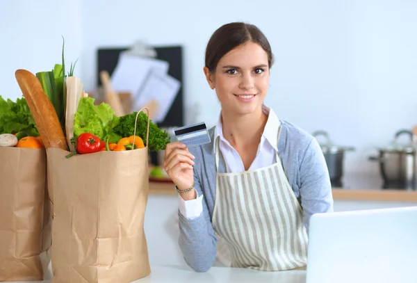Mujer sonriente compras en línea utilizando la computadora y la tarjeta de crédito en la cocina —  Fotos de Stock