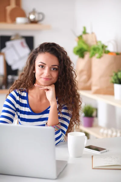 Lachende jonge vrouw met koffiekopje en laptop in de keuken thuis — Stockfoto