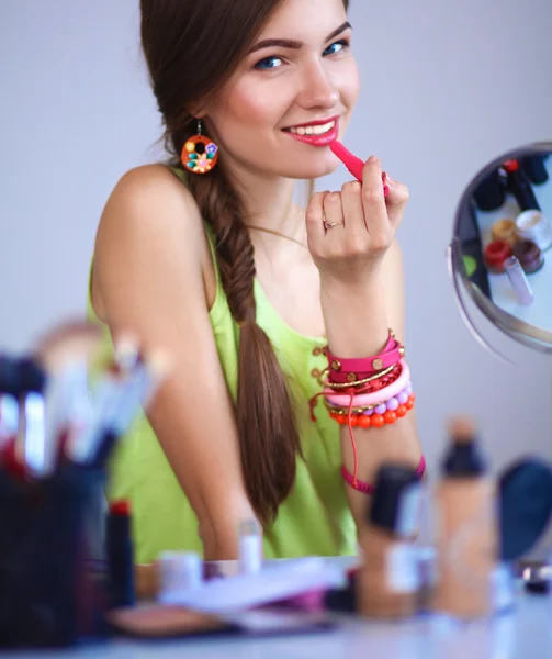 Nuori kaunis nainen tekee meikkiä lähellä peiliä, istuu pöydällä — kuvapankkivalokuva