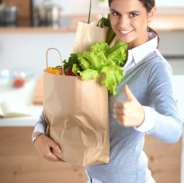 Mujer joven sosteniendo bolsa de la compra de comestibles con verduras. De pie en la cocina —  Fotos de Stock