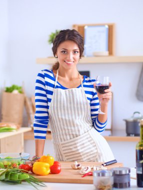 Mutfakta sebze doğrayan, elinde bir bardak şarap tutan genç bir kadın.