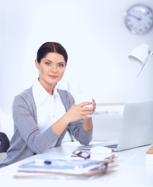 Genç bir iş kadını ofisinde fincanla masada oturuyor.