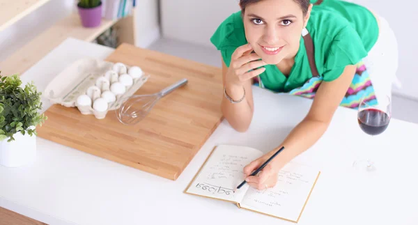 Happy beautiful woman standing in her kitchen writing on a notebook at home — Stock Photo, Image