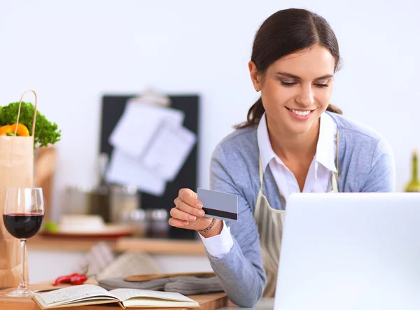 Mujer sonriente compras en línea utilizando la computadora y la tarjeta de crédito en la cocina —  Fotos de Stock