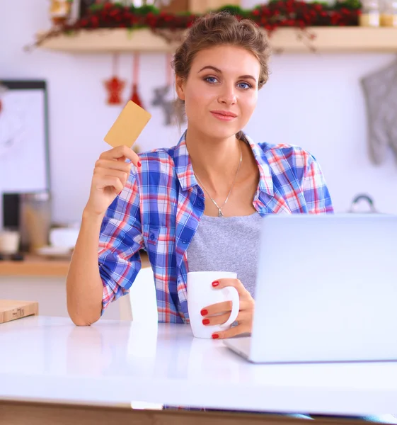 Mujer sonriente compras en línea utilizando la computadora y la tarjeta de crédito en la cocina —  Fotos de Stock