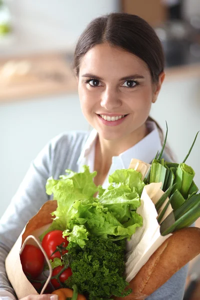 Mujer joven sosteniendo bolsa de la compra de comestibles con verduras. De pie en la cocina —  Fotos de Stock