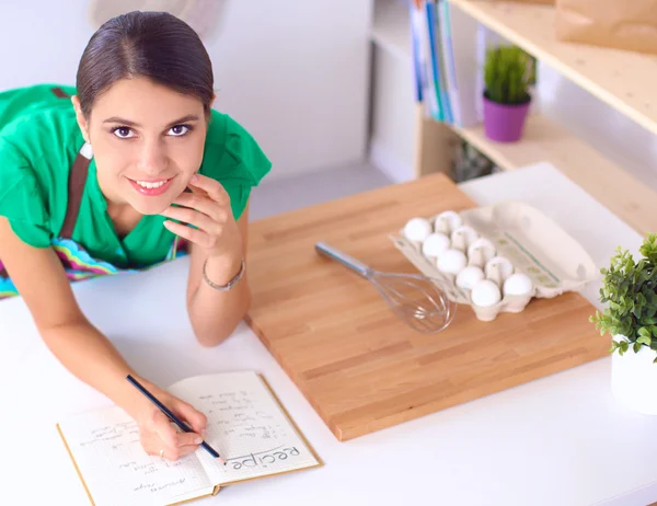 Glückliche schöne Frau steht in ihrer Küche und schreibt zu Hause an einem Notizbuch — Stockfoto