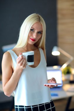 Genç iş kadını elinde fincanla masada oturuyor.