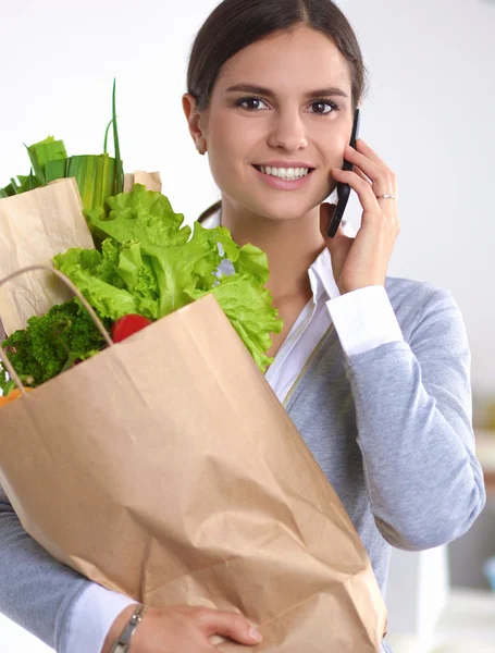 Leende kvinna med mobiltelefon håller shoppingväska i köket — Stockfoto