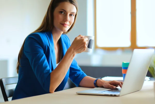 Mulher de negócios sorrindo atraente sentado na mesa do escritório, segurando uma xícara de café . — Fotografia de Stock