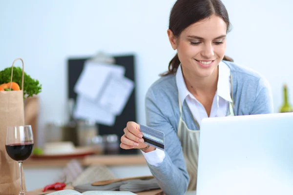 Mujer sonriente compras en línea utilizando la computadora y la tarjeta de crédito en la cocina —  Fotos de Stock