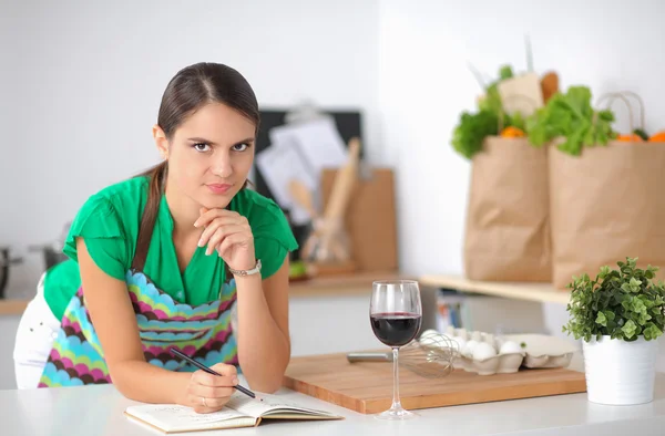 Happy beautiful woman standing in her kitchen writing on a notebook at home — Stock Photo, Image