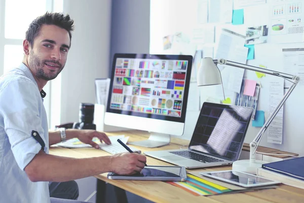 Retrato del joven diseñador sentado en el estudio gráfico frente a la computadora portátil y el ordenador mientras trabaja en línea. — Foto de Stock