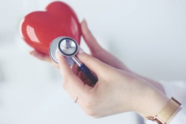 Steteskoplu bir doktor, beyaz arka planda kırmızı kalbi inceliyor.