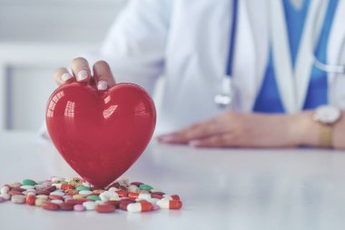 Steteskoplu bir doktor, beyaz arka planda kırmızı kalbi inceliyor.