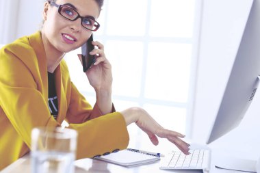 İş kadını ofiste bilgisayar ve cep telefonu kullanarak işine konsantre oluyor.