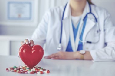 Beyaz önlüklü doktor. Steteskoplu. Elinde haplar ve beyaz bir kalbe takılmış plastik kalp var.