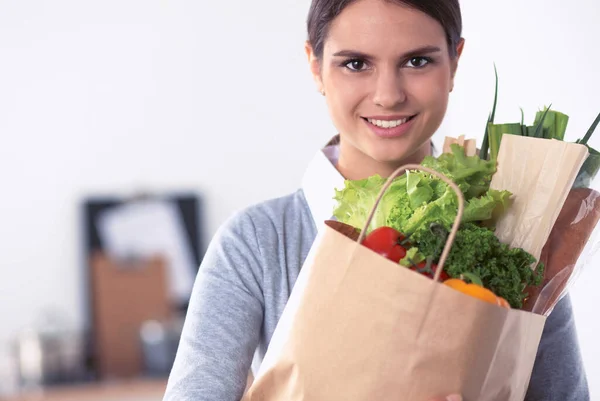 Mujer joven sosteniendo bolsa de la compra de comestibles con verduras de pie en la cocina. — Foto de Stock