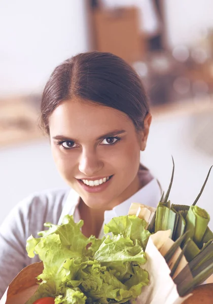 Mujer joven sosteniendo bolsa de la compra de comestibles con verduras de pie en la cocina. — Foto de Stock