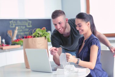 Sebze doğrayan genç bir adam ve mutfakta dizüstü bilgisayarı olan bir kadın.