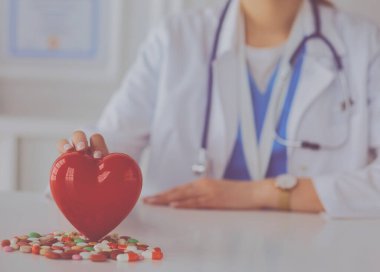 Steteskoplu bir doktor, beyaz arka planda kırmızı kalbi inceliyor.