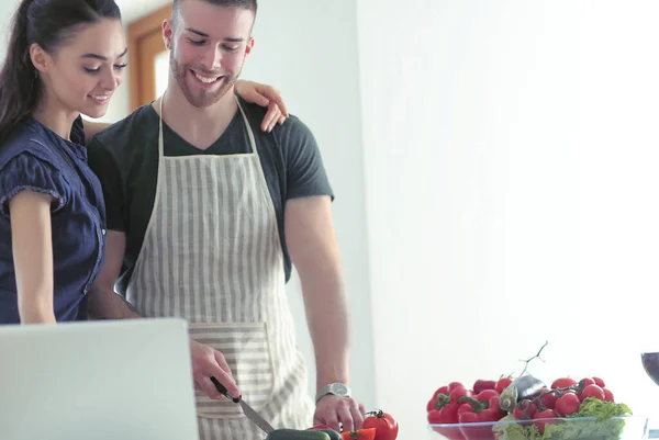 Hombre joven cortando verduras y mujer de pie con el ordenador portátil en la cocina — Foto de Stock