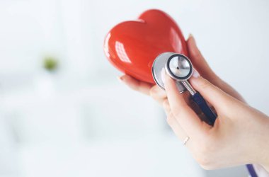 Steteskoplu bir doktor, beyaz arka planda kırmızı kalbi inceliyor.