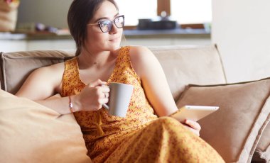 Genç kadın evinde kanepede kahve içerken tablet kullanıyor.