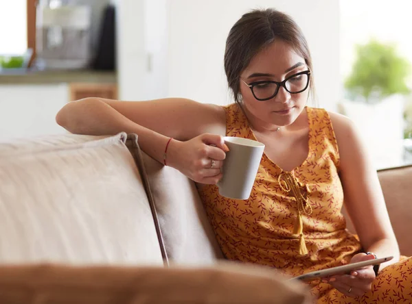Genç kadın evinde kanepede kahve içerken tablet kullanıyor. — Stok fotoğraf