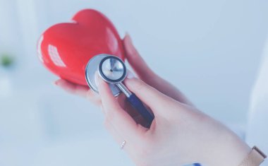 Steteskoplu bir doktor, beyaz arka planda kırmızı kalbi inceliyor.