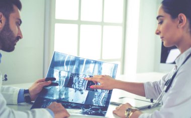 İki doktor röntgen görüntülerini inceliyor, parlak ofiste danışmanlık yapıyorlar.
