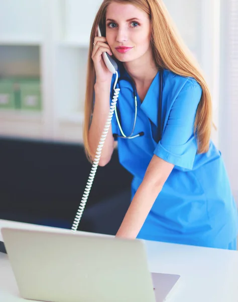 Ung kvinna läkare i vit rock vid datorn med hjälp av telefon — Stockfoto