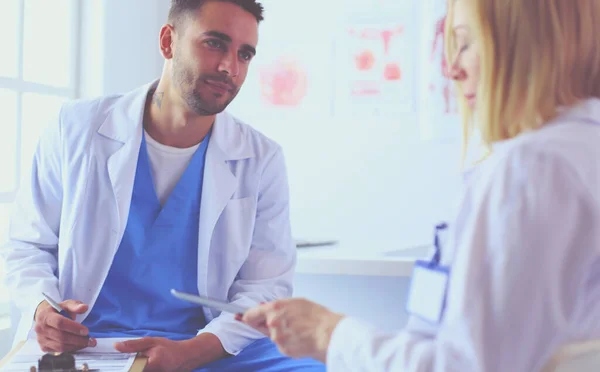 Schöner Arzt spricht mit junger Ärztin und macht sich Notizen, während er in seinem Büro sitzt. — Stockfoto