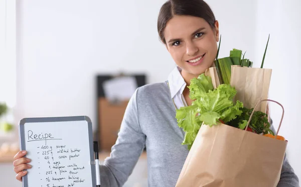 Mujer joven sosteniendo bolsa de la compra de comestibles con verduras de pie en la cocina. — Foto de Stock