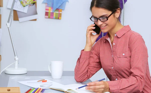 Junge attraktive Modedesignerin arbeitet am Schreibtisch und zeichnet, während sie mit dem Handy spricht — Stockfoto