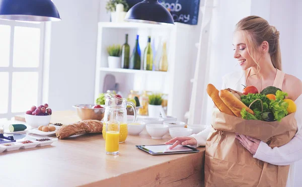 Mujer joven sosteniendo bolsa de la compra de comestibles con verduras. De pie en la cocina —  Fotos de Stock