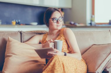 Genç kadın evinde kanepede kahve içerken tablet kullanıyor.