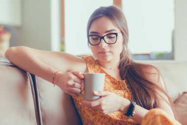 Genç kadın evinde kanepede kahve içerken tablet kullanıyor.