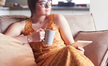 Genç kadın evinde kanepede kahve içerken tablet kullanıyor.