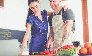 Sebze doğrayan genç bir adam ve mutfakta dizüstü bilgisayarı olan bir kadın.