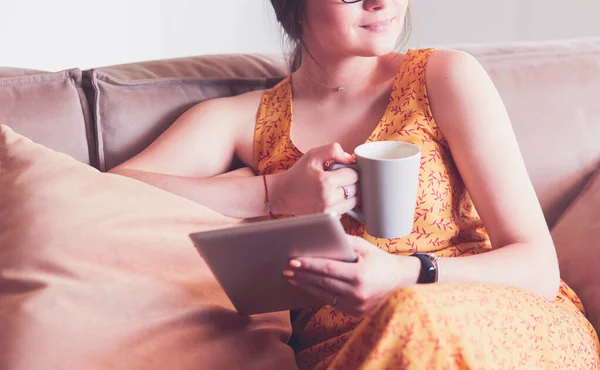 Mujer joven utiliza la tableta mientras se relaja en casa con café en el sofá sofá — Foto de Stock