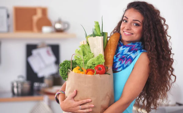 Mujer joven sosteniendo bolsa de la compra de comestibles con verduras de pie en la cocina. — Foto de Stock