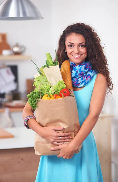 Ung kvinde med indkøbspose med grøntsager stående i køkkenet. - Stock-foto