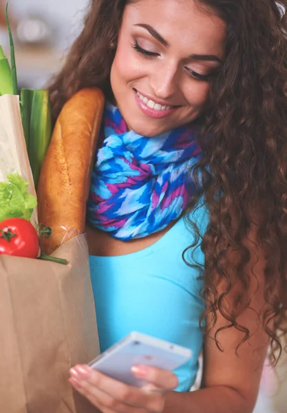 Smiling woman with mobile phone holding shopping bag in kitchen — Stock Photo, Image