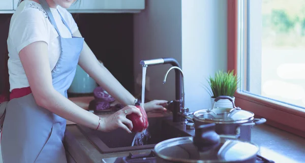Женщина моет овощи. Красивая молодая женщина моет овощи для салата и улыбается стоя на кухне — стоковое фото